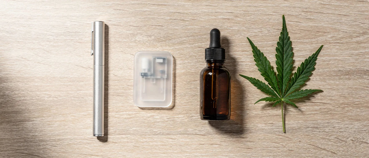 Einweg-Vape oder Pod-System: Stift, Kartusche und Fläschchen nebeneinander auf hellem Holz mit Hanfblatt